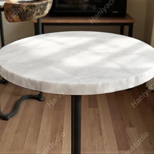 May include: A circular, white marble table top with a natural edge, supported by a black metal pedestal base. The table is set against a blurred background, showcasing a minimalist design.