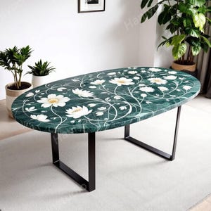 Può includere: Tavolo ovale in marmo verde con motivo floreale bianco e base in metallo nero. Il tavolo è circondato da piante in vasi bianchi e intrecciati. Il tavolo misura circa 152 cm di lunghezza.