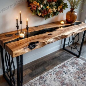 Puede incluir: Una consola de madera con un diseño de borde vivo y una incrustación de resina epoxi negra. La mesa tiene patas de metal negro con detalles decorativos. Hay velas y un jarrón con flores secas sobre la mesa. La mesa mide aproximadamente 152 cm de largo.