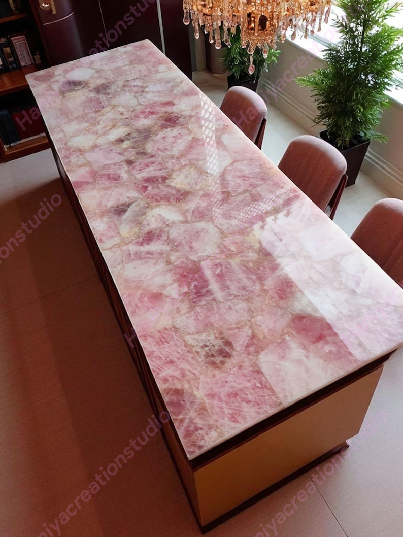 May include: A rectangular table with a rose quartz top. The pink and white stone surface is set on a light brown wooden base. The table is surrounded by chairs and a chandelier.