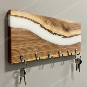May include: Wooden key holder with a natural wood grain design and a white resin river. The key rack has six silver hooks for hanging keys. Two sets of keys are hanging from the hooks. A decorative and functional home decor item.