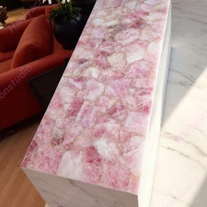 May include: A rectangular, pink rose quartz table with a white marble base. The tabletop is composed of irregular, interlocking pieces of pink quartz. The table is in a well-lit room, with a red sofa and a black vase of white flowers in the background.