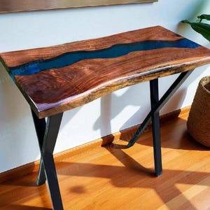 May include: A handcrafted wooden console table with a live edge design. The tabletop features a river of blue epoxy resin flowing through the center. The table is supported by black metal legs. The table is approximately 48 inches long.