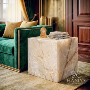May include: A cube-shaped side table made of polished beige and white veined stone. On top sits a small gold-toned analogue clock, a pair of wire-rimmed glasses, and a stack of three hardcover books with visible spines and white pages.