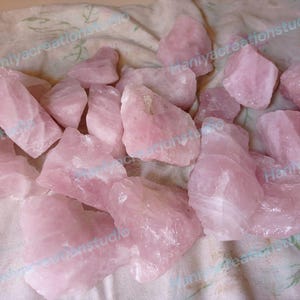 May include: A collection of raw, rough-cut rose quartz crystals. The stones vary in size and shape, displaying a soft pink hue. The crystals are scattered on a light-colored, textured surface, creating a natural and earthy aesthetic.