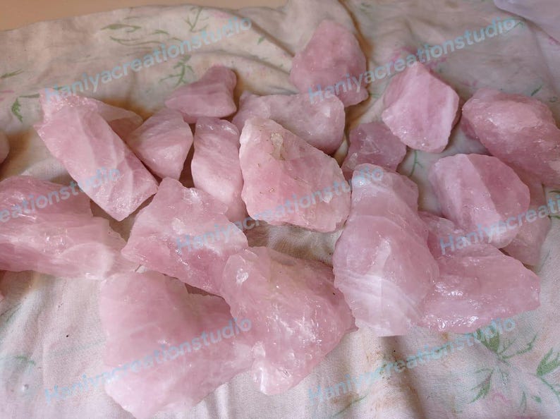 May include: A collection of raw, rough-cut rose quartz crystals. The stones vary in size and shape, displaying a soft pink hue. The crystals are scattered on a light-colored, textured surface, creating a natural and earthy aesthetic.