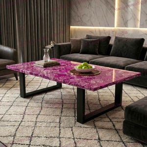 Puede incluir: Una mesa de centro rectangular con una encimera de ágata rosa vibrante y patas de metal negro. La mesa está en un salón, con una alfombra estampada y un sofá gris oscuro en el fondo. Un cuenco de manzanas verdes está sobre la mesa.