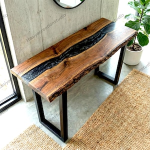 May include: A rectangular console table featuring a natural live-edge wood slab with a prominent central river of translucent black epoxy resin. The table is supported by two black metal U-shaped legs, showcasing a contrast between the wood grain.