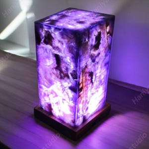 May include: A rectangular lamp with a purple and white marbled design. The lamp is illuminated, showcasing the intricate patterns within. It sits on a dark wooden base, creating a striking contrast. The lamp is a decorative lighting fixture.