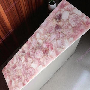 May include: A rectangular table with a top made of pink rose quartz. The quartz is composed of irregular, interlocking pieces in shades of pink and white. The table's base is a light gray color, and the table is positioned against a dark wood wall.