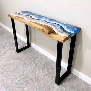 May include: A long, wooden console table with a live edge and a blue and white epoxy resin design resembling ocean waves. The table has black metal legs and is approximately 48 inches long.