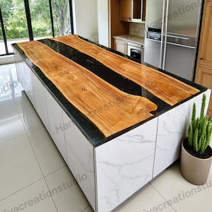 Pode incluir: Ilha de cozinha com bancada de madeira e incrustação de resina epóxi preta. A ilha tem uma base branca com padrão de mármore e portas de armário. Uma planta em vaso está na lateral. A ilha está numa cozinha moderna. A bancada tem cerca de 240 cm.
