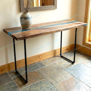 May include: A rectangular console table featuring a natural wood grain top with a central blue resin inlay. The table is supported by four black metal pipe legs arranged in two U-shaped bases. A ceramic vase sits on the left side of the table surface.