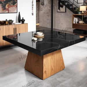 Może przedstawiać: Nowoczesny kwadratowy stół jadalniany z czarnym marmurowym blatem i jasnobrązową drewnianą podstawą. Stół ma minimalistyczny design i znajduje się w nowoczesnej jadalni. Na stole stoi mała taca z filiżanką i spodkiem.