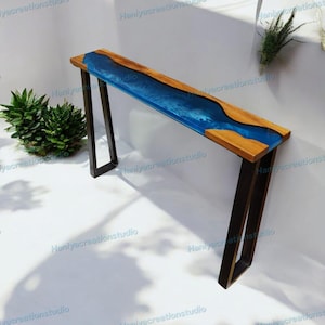 Puede incluir: Una consola larga y estrecha con una tapa de madera y un diseño de río de resina azul. La mesa tiene patas de metal oscuro y mide aproximadamente 122 cm de largo. La madera tiene una veta natural y la resina tiene un acabado brillante.