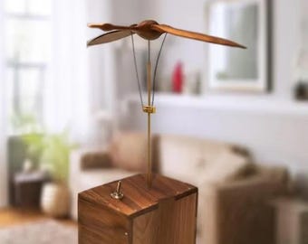 May include: A wooden kinetic sculpture featuring a dragonfly design. The sculpture has a wooden box base with a brass rod supporting the dragonfly. The dragonfly's wings are made of wood and are positioned to appear as if they are in flight. The sculpture is a decorative item.