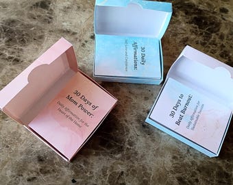 May include: Three open boxes with cards inside, each with a different title. The boxes are pink and blue, and the cards have titles such as "30 Days of Mom Power" and "30 Days to Beat Burnout."