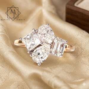 Può includere: Un anello d'oro con un gruppo di pietre preziose chiare in tagli smeraldo, a goccia e quadrati. L'anello è esposto su un tessuto beige. Il marchio "MANIYA JEWELS" è visibile.