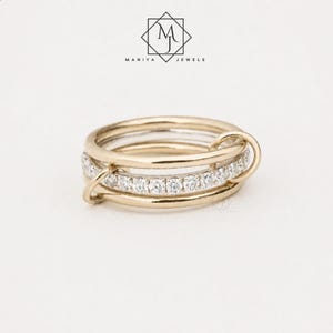 May include: A gold ring featuring a central band encrusted with a row of clear, round-cut stones. The central band is encircled by two smooth, plain gold bands that are linked together by a small gold loop. The logo Maniya Jewels is visible.
