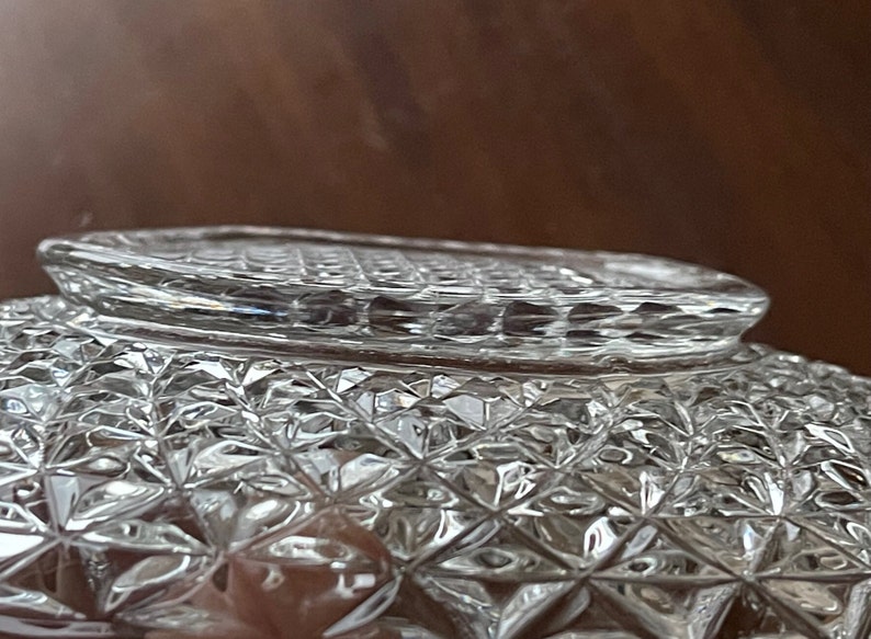 May include: Close-up of a clear, cut-glass bowl with a diamond pattern. The bowl's rim has a textured design. The glass reflects light, creating a sparkling effect. The bowl is likely for serving or display.