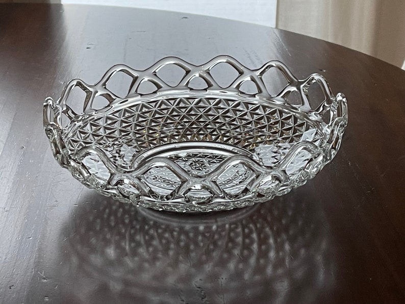 May include: A clear, oval-shaped glass serving bowl with a diamond-cut pattern and a decorative openwork rim. The bowl is designed with a scalloped edge and sits on a dark wooden surface. This elegant piece is suitable for serving fruit or decorative purposes.