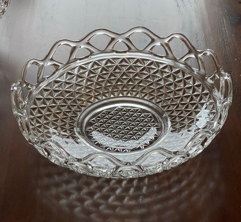 May include: Clear glass serving dish with a diamond pattern and a decorative, openwork rim. The dish has an oval center and a scalloped edge, perfect for serving appetizers or desserts. The glass reflects light, creating a sparkling effect.