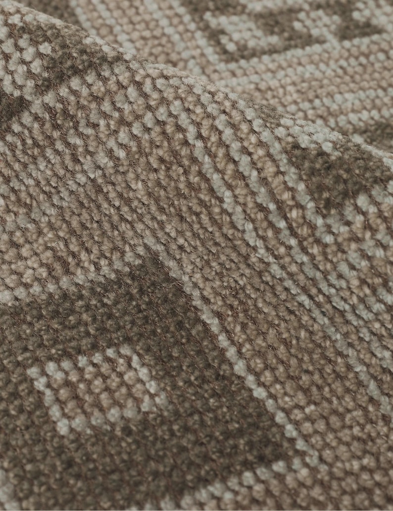 May include: Close-up of a textured, woven fabric in shades of brown and beige. The fabric has a repeating pattern of geometric shapes and lines, creating a subtle, yet intricate design. The material appears soft and slightly folded.