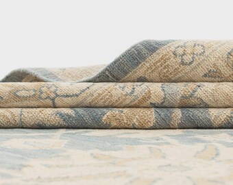 May include: Close-up of a rolled-up area rug with a floral pattern. The rug features a beige and light blue color scheme. The edges are a darker blue, and the rug appears to be made of a woven material.