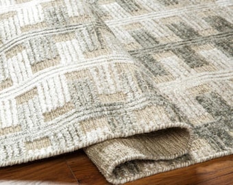 Puede incluir: Una alfombra enrollada con un patrón geométrico en tonos beige, gris y blanco. La textura de la alfombra es visible y está sobre un suelo de madera. El diseño presenta formas rectangulares.
