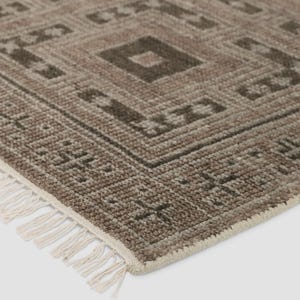 May include: A close-up of a rectangular rug with a geometric pattern in shades of brown and beige. The rug features a repeating diamond and square design. The edge of the rug has a fringe of white tassels.
