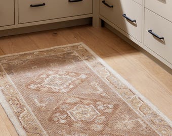 Puede incluir: Una alfombra rectangular de aspecto envejecido, con un diseño central de medallón en tonos marrón, beige y crema. La alfombra está colocada sobre un suelo de madera clara, con armarios blanquecinos y tiradores negros al fondo.