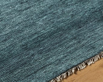 May include: Close-up of a teal rug with a textured surface and a fringe along one edge. The rug is placed on a wooden floor. The rug's colour varies slightly, creating a subtle visual texture.
