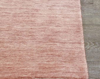 Puede incluir: Primer plano de una alfombra rectangular de color rosa descolorido. La alfombra tiene una superficie texturizada y un fleco en un borde. Está colocada sobre un suelo de madera. Las fibras de la alfombra tienen un aspecto sutil y moteado.