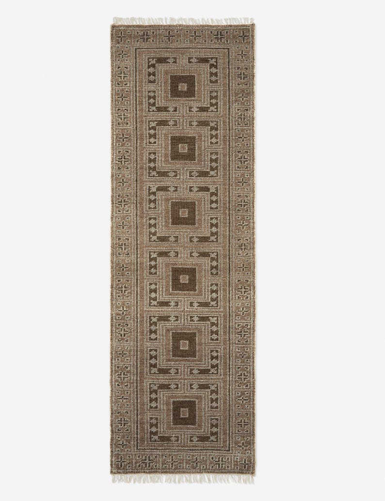 May include: A rectangular brown and beige patterned rug with a repeating geometric design. The rug features a series of squares within squares, creating a symmetrical pattern. The edges are finished with a fringe.