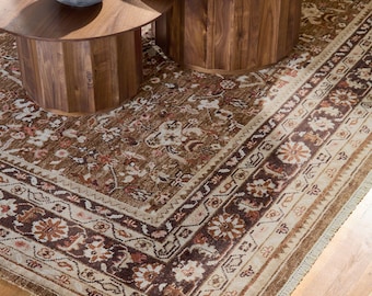 Puede incluir: Una alfombra estampada marrón y beige con un borde detallado. La alfombra presenta diseños florales y geométricos en tonos marrones, beige y rojos. La alfombra está colocada sobre un suelo de madera, con dos mesas de centro de madera al fondo.