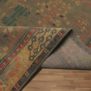 May include: An olive green rug with a folded corner, revealing a patterned underside. The rug features a detailed design with teal, red, and gold accents. The border has a repeating floral pattern.