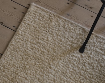 Puede incluir: Primer plano de una alfombra texturizada de color crema con un patrón tejido. La alfombra es rectangular y parece estar hecha de una fibra natural gruesa. Un objeto de metal negro es parcialmente visible en la esquina superior derecha. La alfombra está sobre un suelo de madera clara.