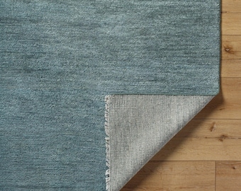 May include: A teal rug with a textured surface. The corner is folded back, showing a lighter grey underside with a fringed edge. The rug is displayed on a wooden floor.
