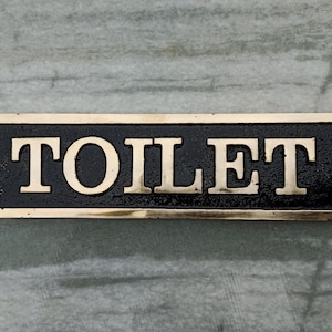 Könnte beinhalten: Rechteckiges schwarzes und goldenes Toilettenschild. Das Wort "TOILET" ist in goldfarbenen Buchstaben geschrieben. Das Schild hat einen goldenen Rand und ist auf einer grau-weißen marmorähnlichen Oberfläche montiert.