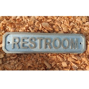 Könnte beinhalten: Ein rechteckiges, hellblaues Metallschild mit dem Wort "RESTROOM" in erhabenen, silbernen Buchstaben. Das Schild ist vor einem Hintergrund aus braunen Holzspänen angebracht. Das Schild hat abgerundete Ecken und eine Vintage-Ästhetik.