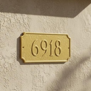 May include: A gold-coloured rectangular address plaque displaying the number "6918" in a serif font. The plaque is mounted on a textured, light-coloured stucco wall, with a terracotta tile roof above.
