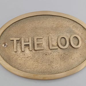 May include: An oval brass plaque with the words "THE LOO" in raised lettering. The sign has a textured surface and two small holes for mounting. A decorative sign for a toilet or restroom.