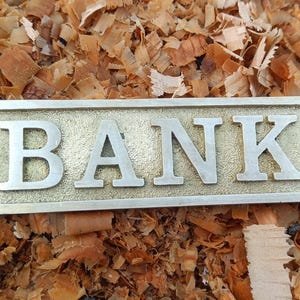 Könnte beinhalten: Ein rechteckiges Messingschild mit dem Wort "BANK" in erhabenen silbernen Buchstaben. Das Schild hat zwei Löcher zur Befestigung und liegt auf einem Hintergrund aus Holzspänen.