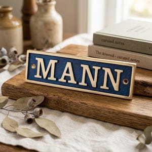 May include: A rectangular metal sign with a dark blue textured background and raised gold-colored lettering that spells out the word MANN. The sign features a gold-colored border and two circular mounting holes, one on each far left and right side.