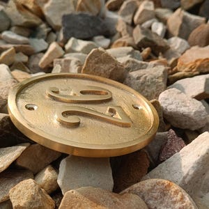May include: A circular brass house number plaque featuring the embossed number 26 in the center. The plaque has a smooth, metallic finish with two small pre-drilled holes on the left and right sides for mounting purposes, resting on a bed of stones.