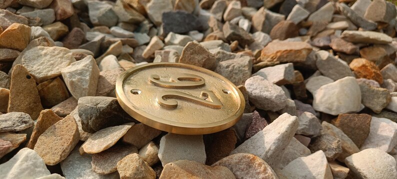 May include: A circular brass house number plaque featuring the embossed number 26 in the center. The plaque has a smooth, metallic finish with two small pre-drilled holes on the left and right sides for mounting purposes, resting on a bed of stones.