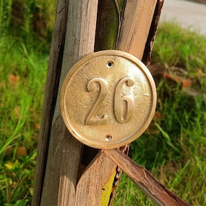 May include: A circular brass house number plaque featuring the embossed number 26 in the center. The plaque has a metallic gold finish with a slightly weathered texture and two small circular mounting holes at the top and bottom edges.