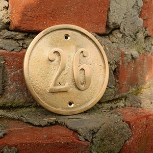 May include: A circular brass house number plaque featuring the embossed number 26 in the center. The plaque has a smooth, metallic finish with a raised border and two small circular mounting holes positioned at the top and bottom of the disc.