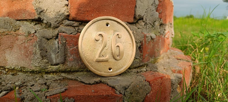 May include: A circular brass house number plaque featuring the embossed number 26 in the center. The plaque has a smooth, metallic finish with a raised border and two small circular mounting holes positioned at the top and bottom of the disc.