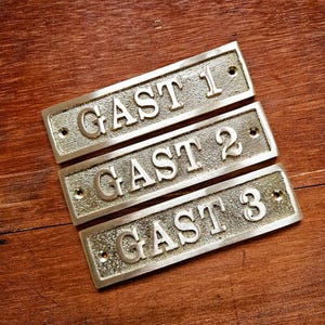 Könnte beinhalten: Drei rechteckige Messingplatten mit dem Wort "GAST" und den Zahlen 1, 2 und 3. Die Platten sind auf einer Holzoberfläche montiert. Jede Platte hat zwei Schraubenlöcher.